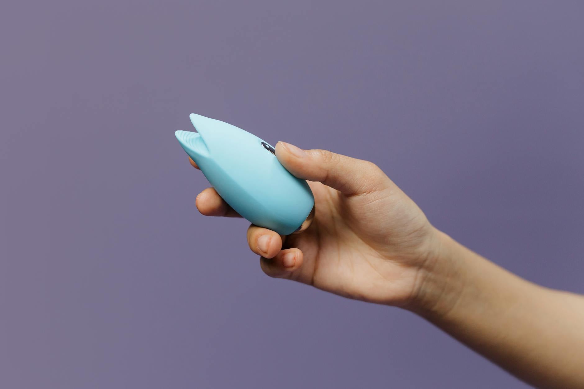 A blue clitoral vibrator held in hand against a solid purple background